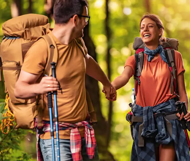 Šťastný pár na túre v lese, nesúci batohy a držiaci sa za ruky, užívajúci si prírodu a outdoorové dobrodružstvo.